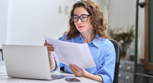 Woman reading a paper while sitting at a laptop